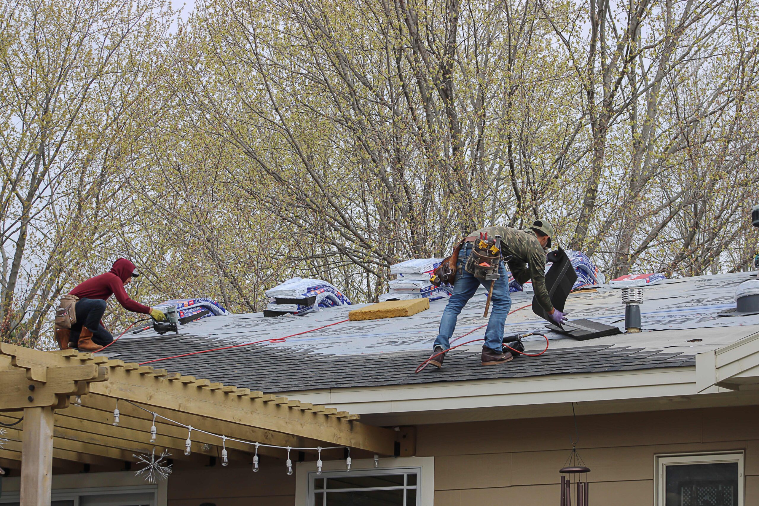 Two workers are installing shingles on a house roof, surrounded by roofing materials and tools. A stucco contractor Twin Cities team could also assist with storm damage stucco repair to fully restore your home’s exterior. Trees line the background.