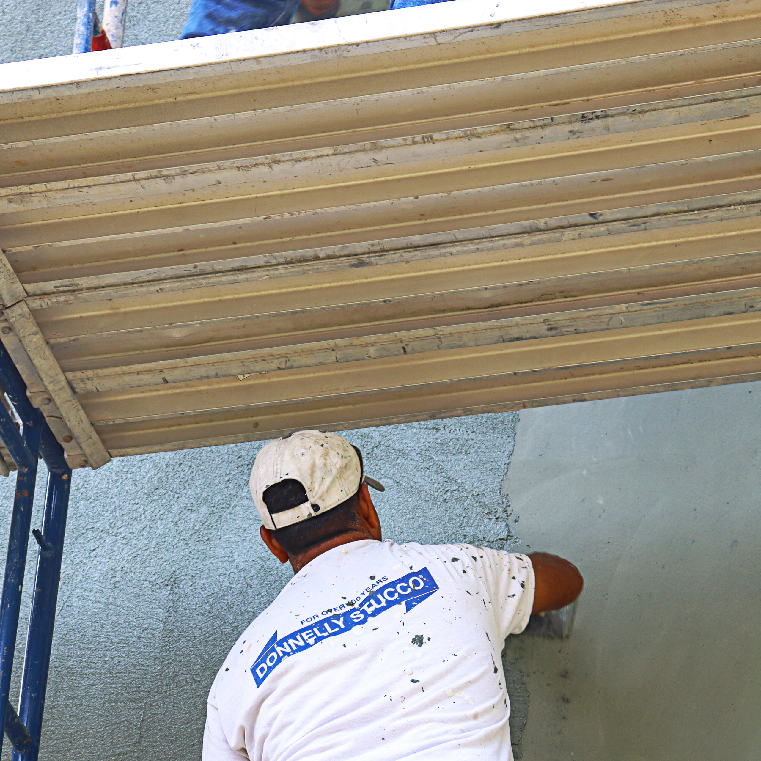 A construction worker in a white shirt and cap applies stucco to a wall beneath a metal scaffold, showcasing expert stucco repair Minnesota services.