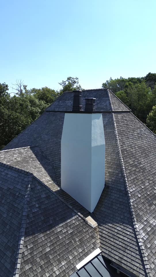 A white chimney with two black caps rises from the center of a multi-faceted shingled roof, expertly restored by an exterior contractor Minnesota, surrounded by green trees under a clear blue sky.