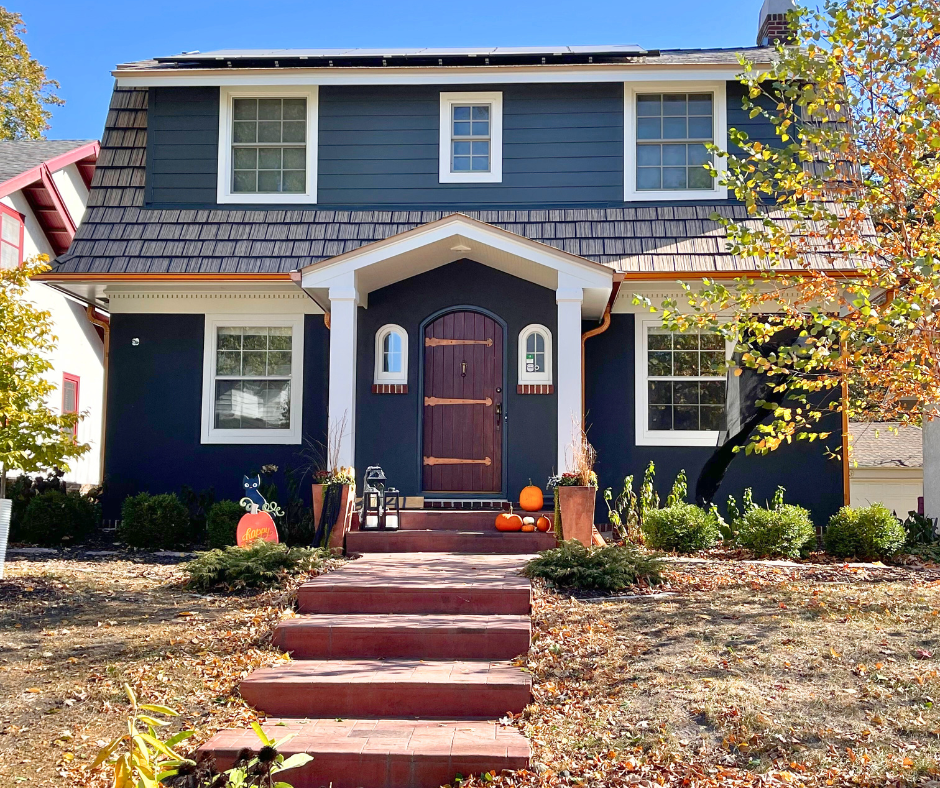 Two-story dark blue house with a wooden front door, white trim, and autumn decorations, including pumpkins and potted plants, on the front porch—expertly maintained by an exterior contractor Minnesota trusts.