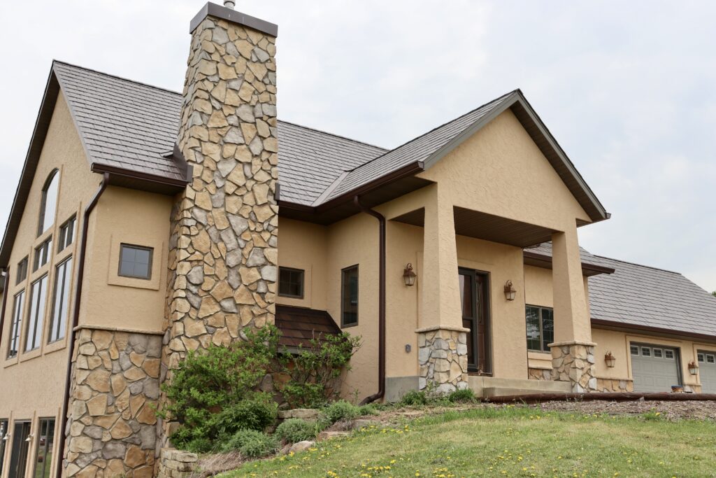 A beige two-story house with large stone chimneys, tall windows, a sloped roof, and a three-car garage on a grassy yard—ideal for anyone seeking expert stucco repair Minnesota services after storm damage.