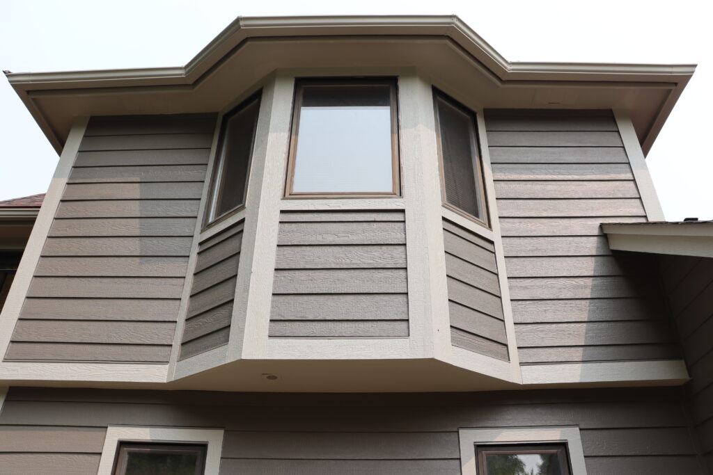 Close-up view of a house’s bay window with beige trim and brown horizontal siding, photographed from below—a typical setting for stucco crack repair or storm damage stucco repair in Minnesota homes.