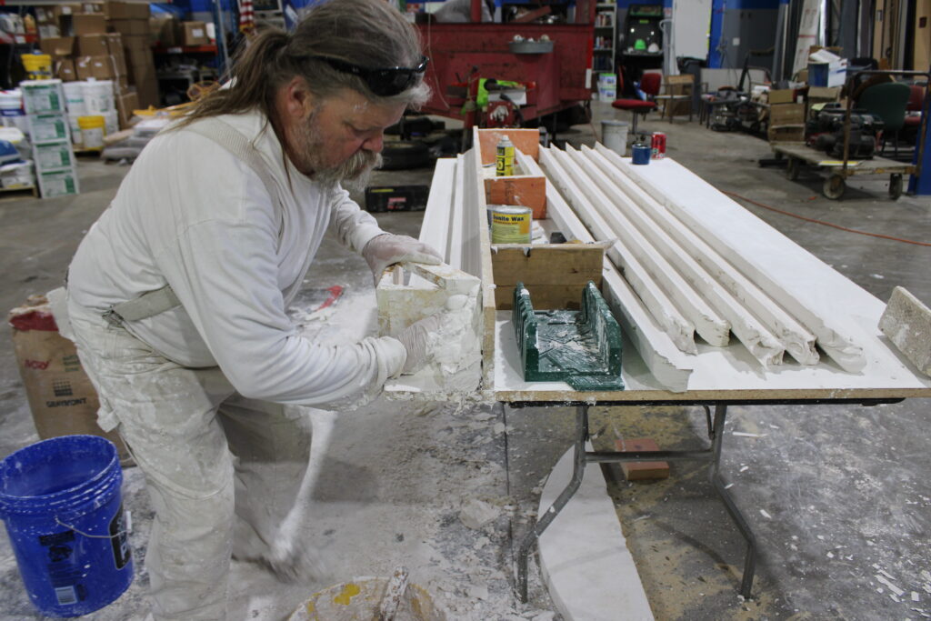 A man wearing work clothes shapes plaster using a mold at a workshop table covered with plaster and long decorative trim pieces, demonstrating skills essential for stucco repair Minnesota projects.