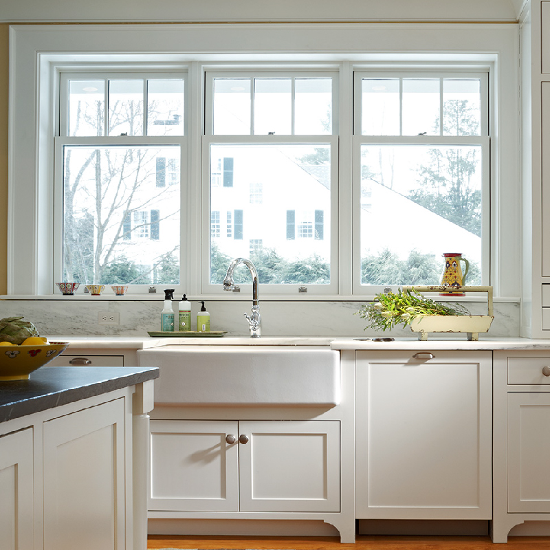 A bright kitchen with white cabinets, a farmhouse sink, a window with a view outside, and various items including soap, dishes, and fresh greens on the counter—perfect for homeowners seeking stucco repair Minnesota.