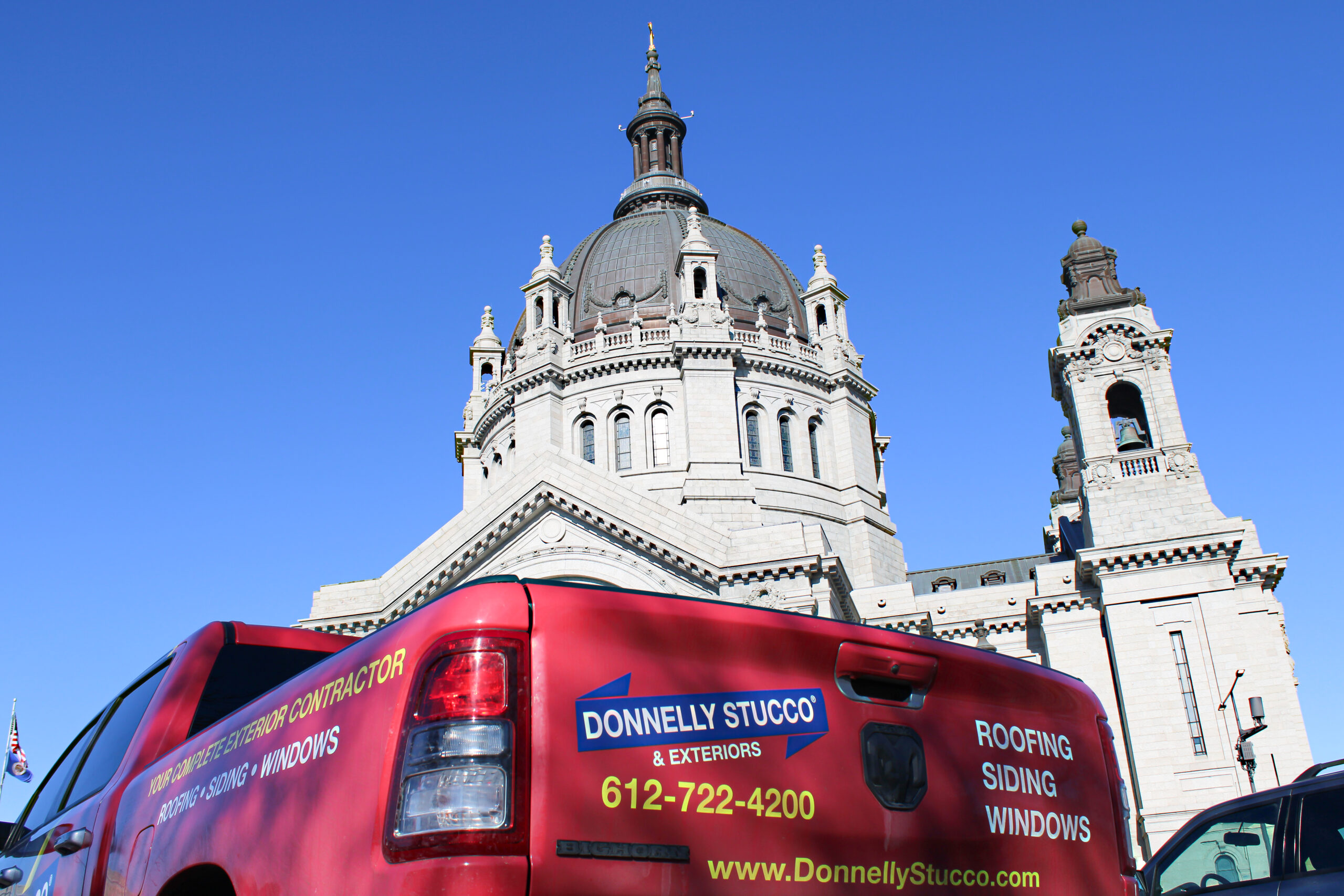 A red Donnelly Stucco & Exteriors truck, trusted stucco contractor Twin Cities, is parked in front of a large stone cathedral with a dome and tower under a clear blue sky.