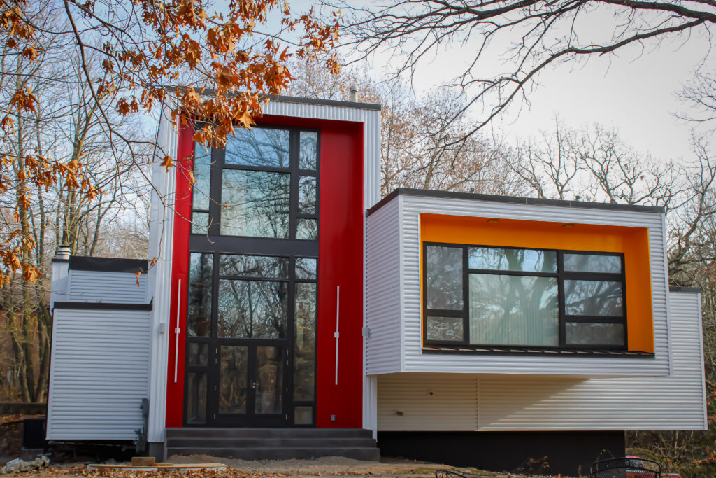 A modern house with large glass windows, a prominent red vertical accent, and a cantilevered section featuring an orange window frame stands among bare trees—ideal for those seeking stucco repair Minnesota or moisture remediation stucco services.