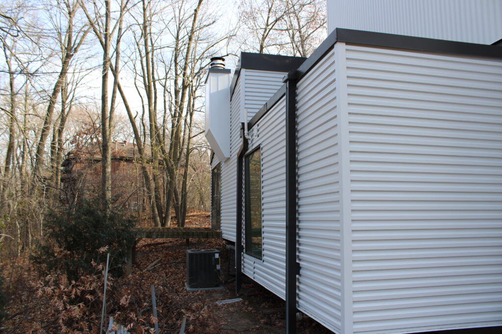 A modern white corrugated metal building stands beside a wooded area with bare trees and fallen leaves, highlighting the importance of moisture remediation stucco for nearby structures in Minnesota’s challenging climate.