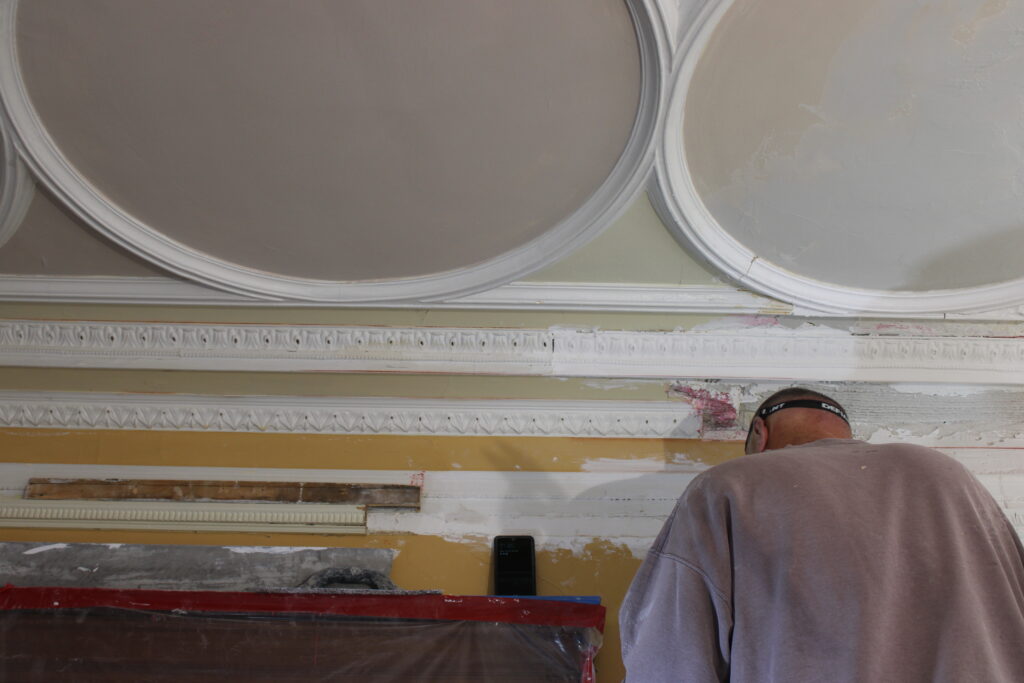 A person wearing a brown shirt stands beneath an ornate, partially painted ceiling and wall, appearing to work on restoration or stucco repair Minnesota.