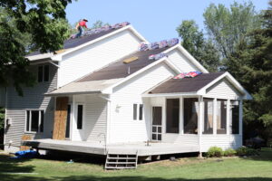A person works on the roof of a white house, installing roofing materials. Bundles of shingles are placed on the roof, and part of the house is covered with roofing felt—ideal for storm damage stucco repair or moisture remediation stucco services.