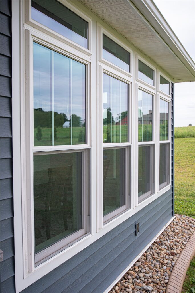 A row of white-framed double-hung windows on a blue house, with a stone border at the base—beautiful work by an exterior contractor in Minnesota, set against a grassy field in the background.