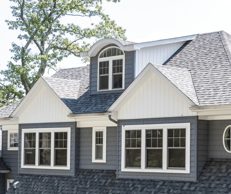 The image shows the upper portion of a gray house with white trim and multiple windows, featuring gabled and arched rooflines—ideal for an exterior contractor Minnesota specializing in stucco crack repair or storm damage stucco repair.