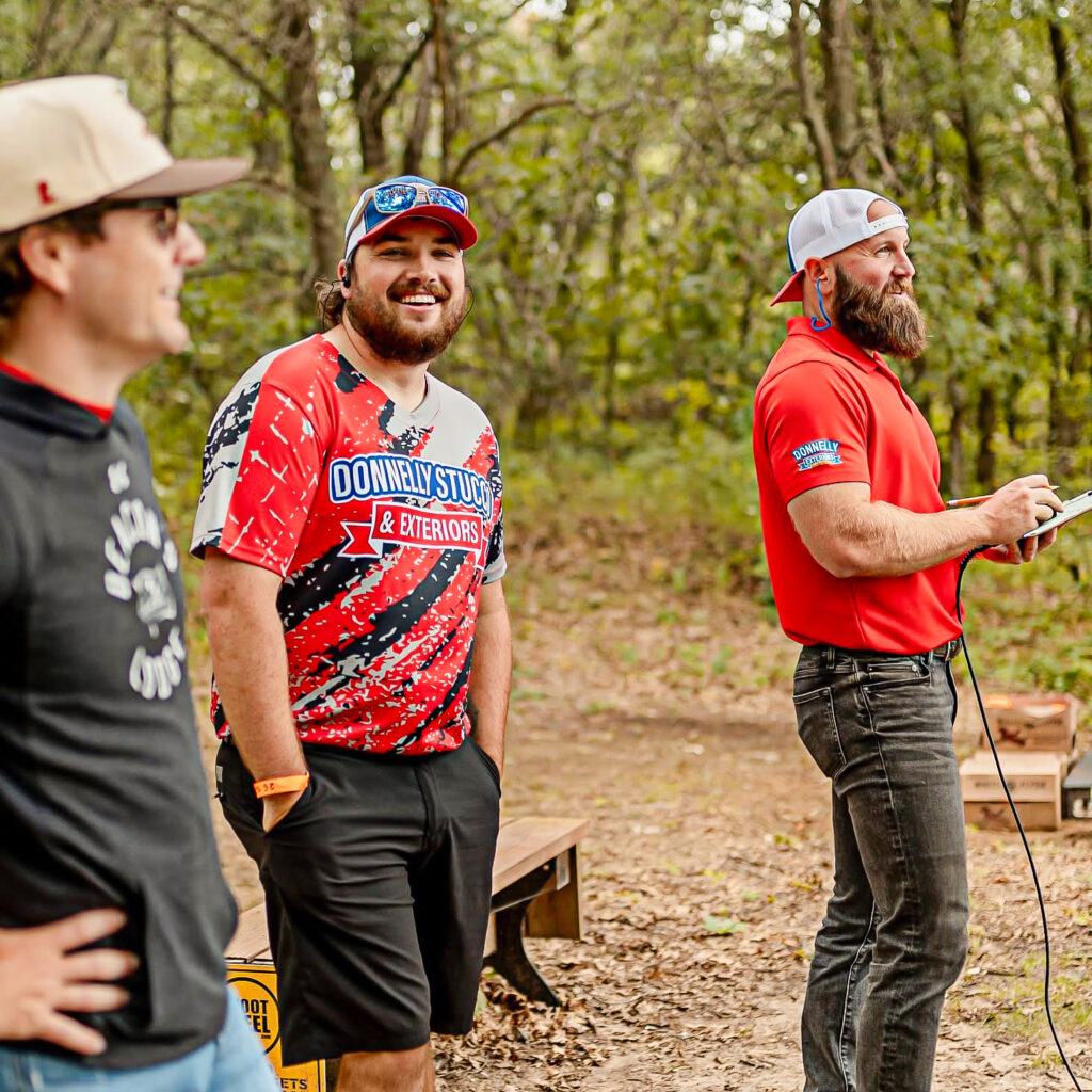 Three men stand outdoors in a wooded area, casually dressed, with one holding a clipboard and pen. Two men are smiling as they discuss moisture remediation stucco solutions, creating a relaxed and informal atmosphere.
