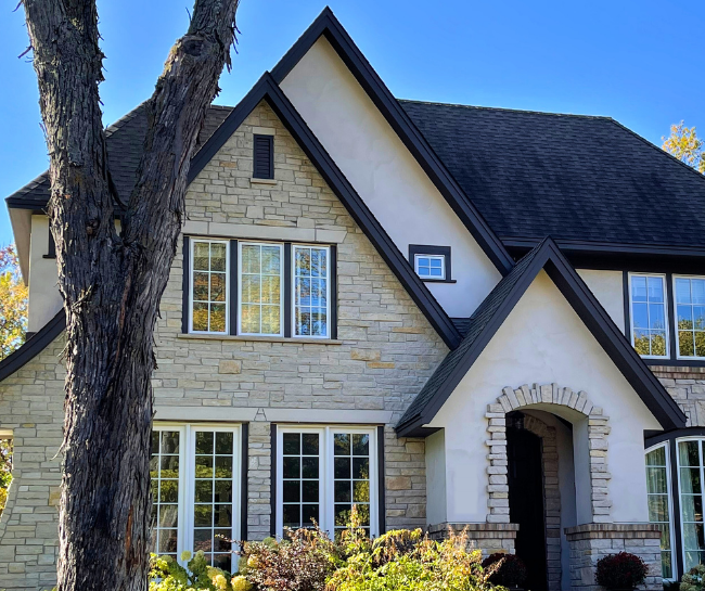 Two-story house with beige stone and stucco exterior, black roof, multiple windows, arched entryway, and a tree in the foreground—ideal inspiration if you’re seeking a trusted stucco contractor Twin Cities for expert craftsmanship.