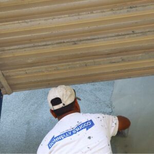 A worker in a white "Donnelly Stucco" t-shirt applies stucco to a wall under a partially closed metal shutter.