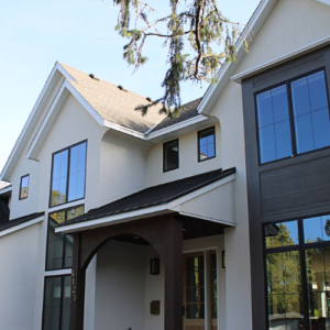 Modern two-story house with large black-trimmed windows, beige exterior walls, and a covered front entrance. A tree branch is visible above the roofline.