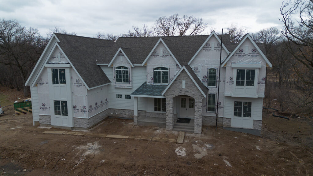 Two-story house under construction with partially finished exterior, stone accents, and construction materials visible around the site.