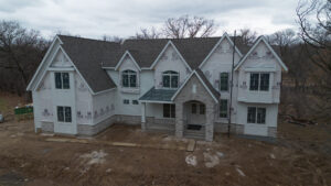 Two-story house under construction with partially finished exterior, stone accents, and construction materials visible around the site.