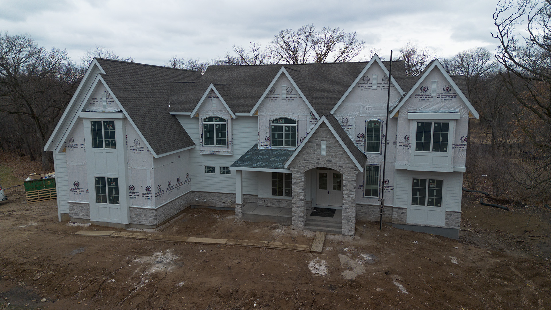 Two-story house under construction with partially finished exterior, stone accents, and construction materials visible around the site.