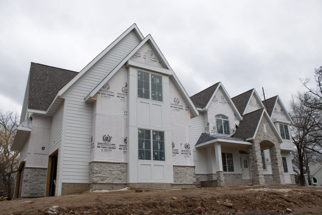 A large, partially constructed house with white siding, stone accents, and exposed building wrap under a cloudy sky.