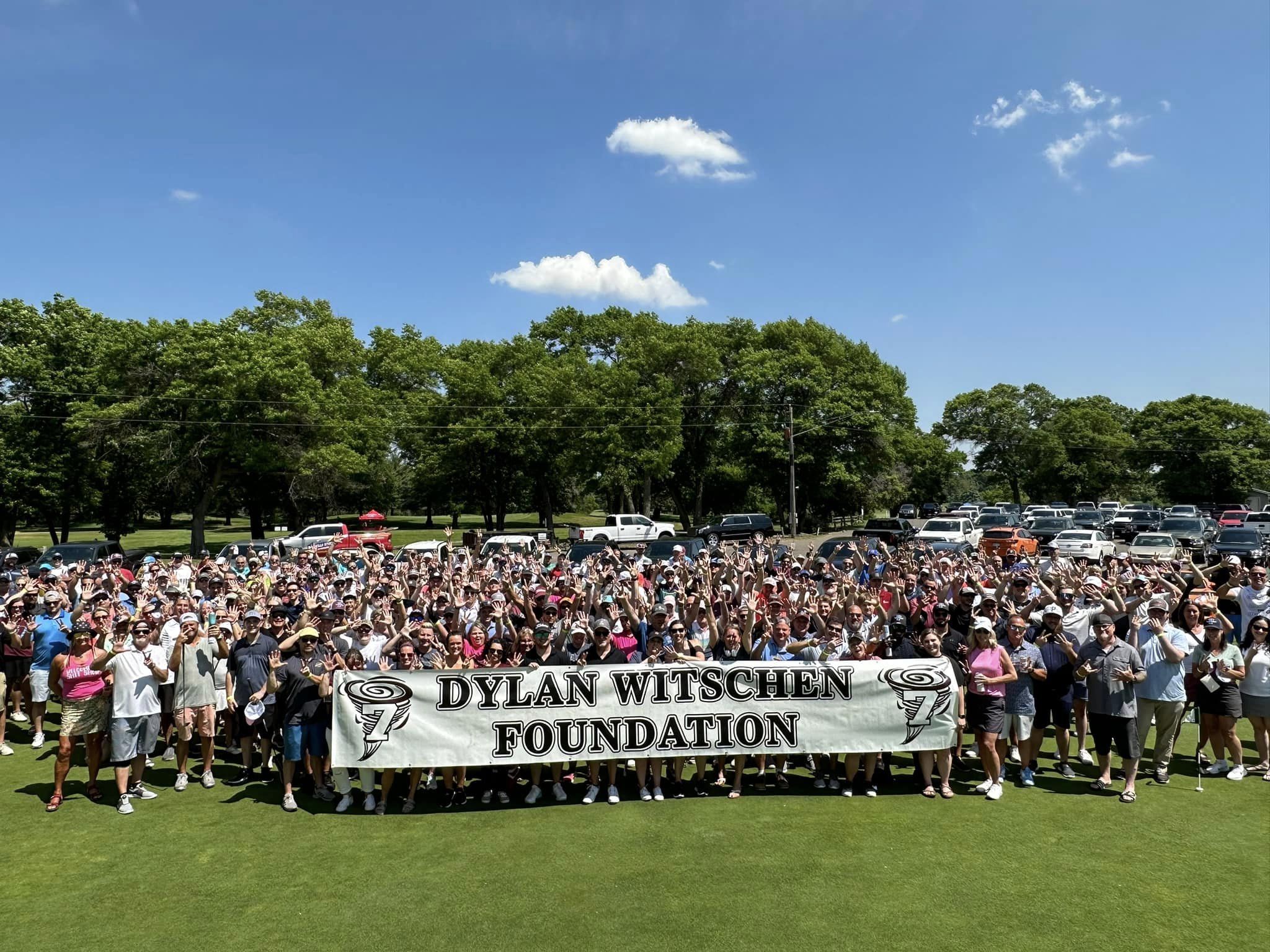 Large group of people outdoors on grass, holding a banner that reads "Dylan Witschen Foundation," with trees and parked cars in the background under a clear blue sky.