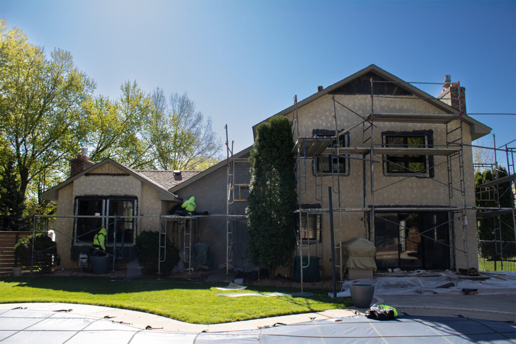 A two-story house under renovation with scaffolding, construction materials, and workers applying stucco to the exterior walls on a sunny day.