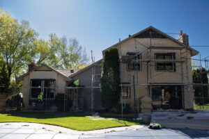 A two-story house under renovation with scaffolding, construction materials, and workers applying stucco to the exterior walls on a sunny day.