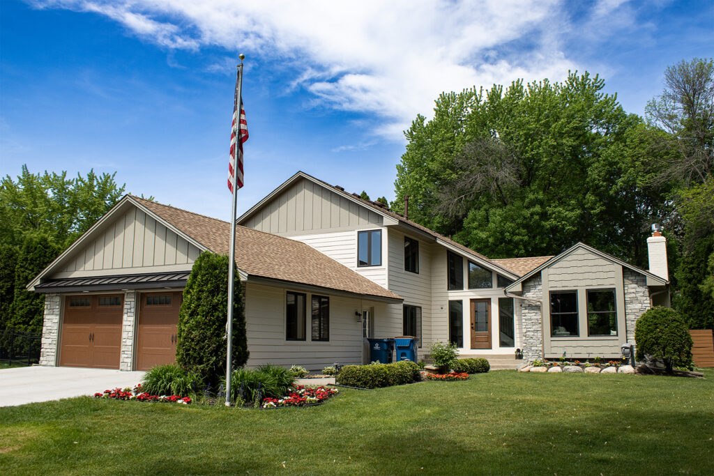Two-story suburban house with a well-kept lawn, two-car garage, flower beds, and an American flag on a pole in the front yard, set against a backdrop of trees and blue sky.