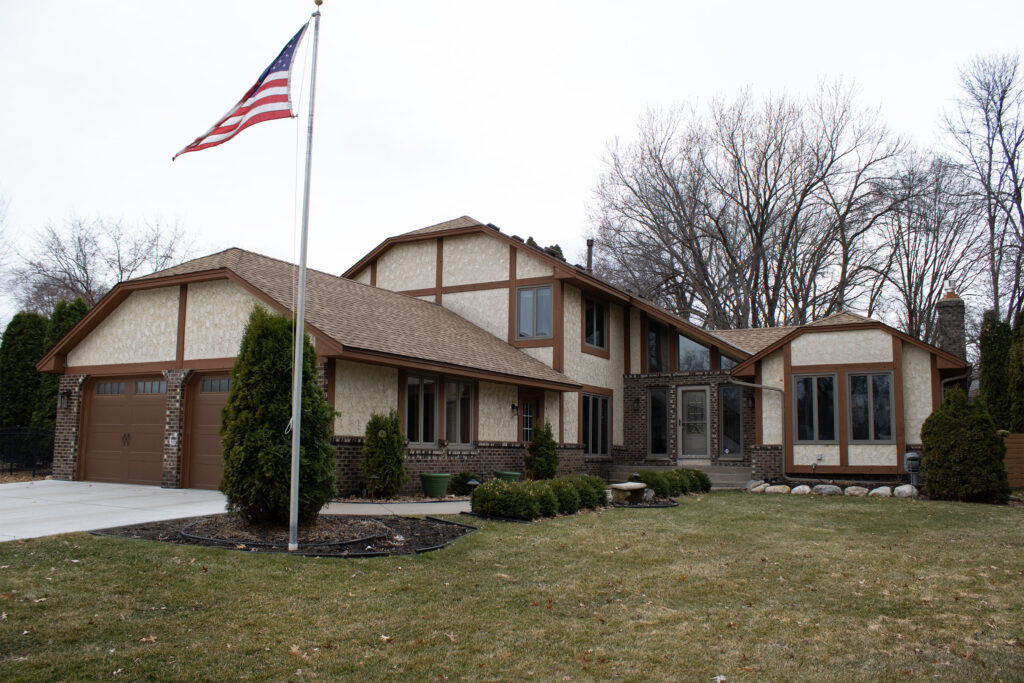 Two-story suburban house with brown trim, attached garage, and manicured lawn; an American flag is raised on a flagpole in the front yard.