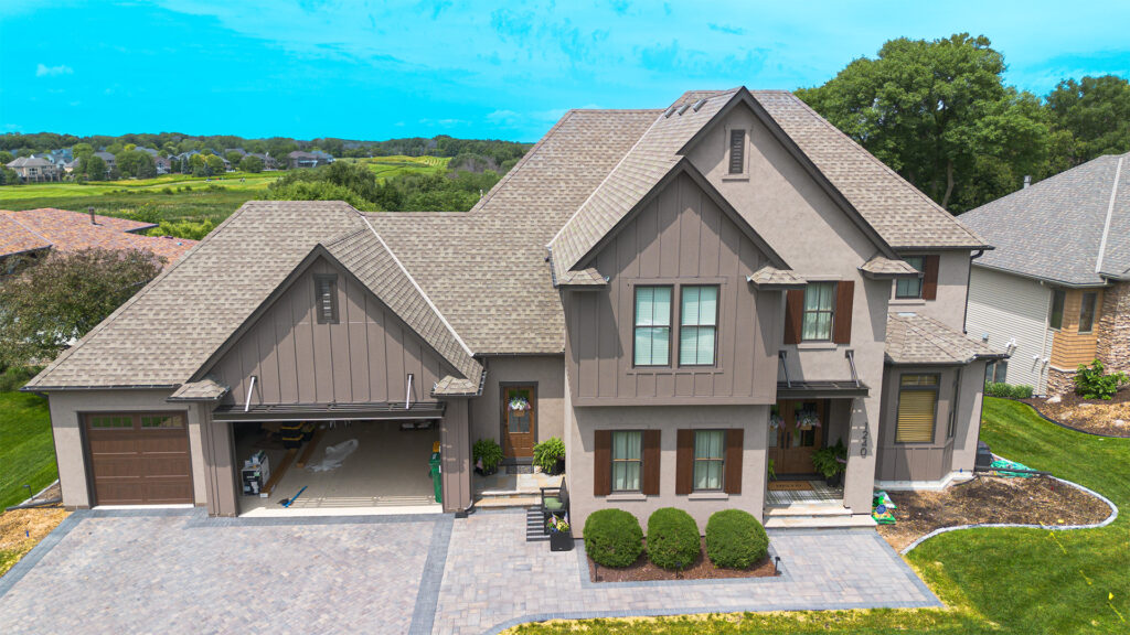 A two-story suburban house with a double garage, brown siding, manicured bushes, and a paved driveway, surrounded by green lawns and neighboring houses.