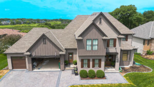 A two-story suburban house with a double garage, brown siding, manicured bushes, and a paved driveway, surrounded by green lawns and neighboring houses.